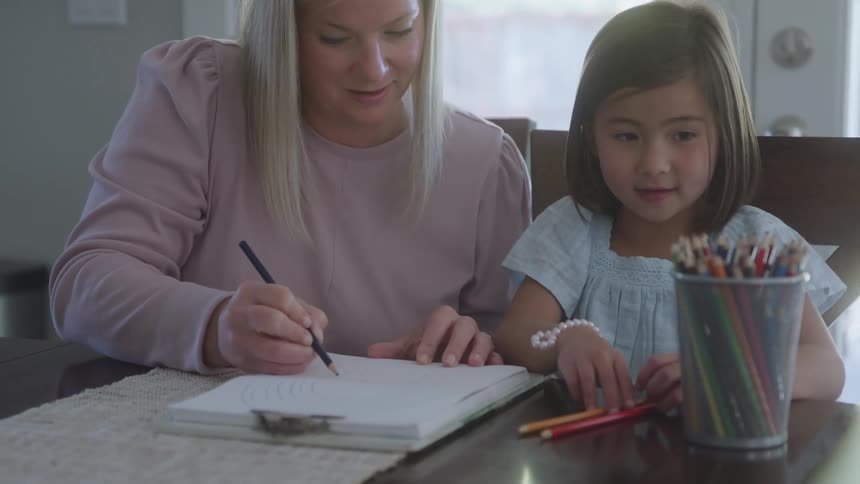 A woman in a pink sweater draws a spiral with a black pencil in a sketchbook as a young girl in a light blue dress watches and reaches for colored pencils nearby on a wooden table