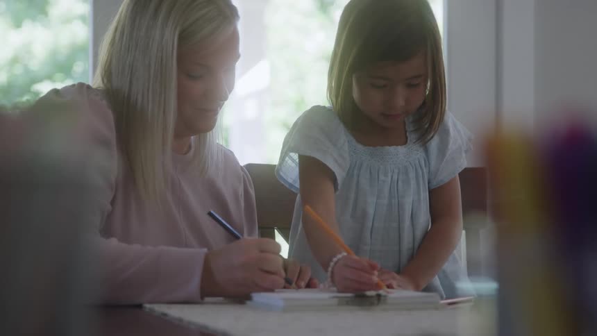 Mother and daughter drawing and colouring with pencils in a book placed on a table indoors during daytime