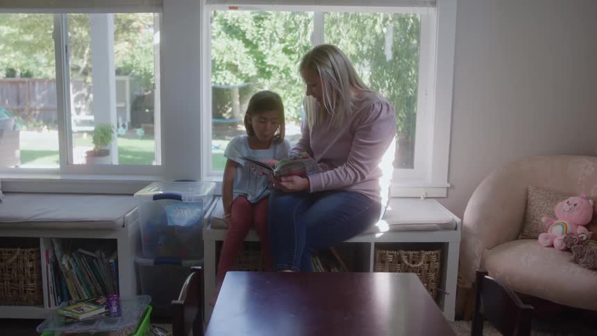 A woman in a pink sweater and jeans sits on a window seat next to a young girl in a light blue dress as they read a book together in a brightly lit room overlooking a sunny backyard