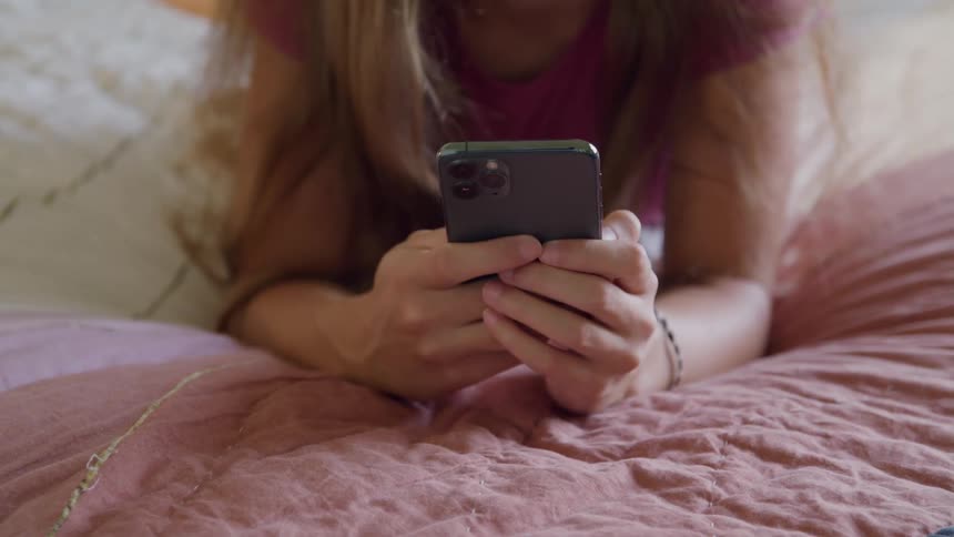 White female lying down in bed texting on a smartphone indoors during day time