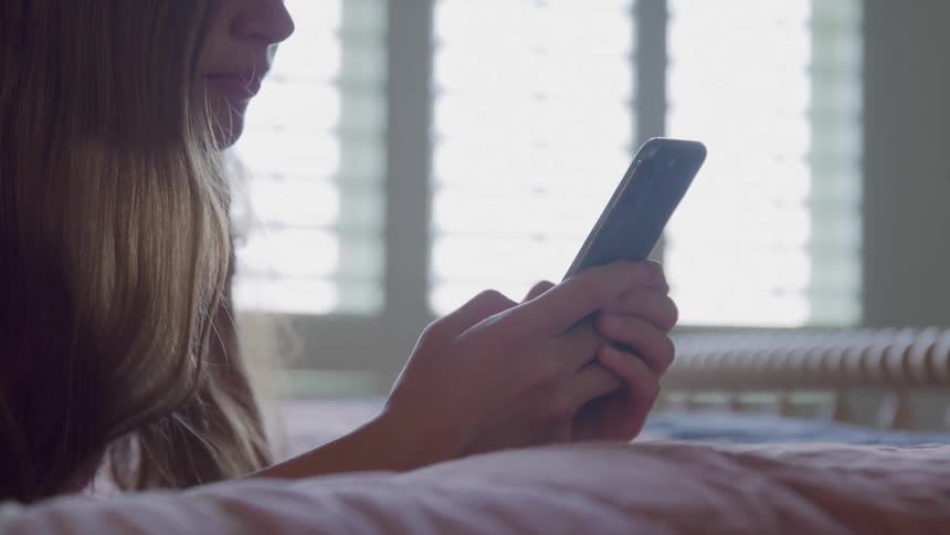 A girl with long brown hair lies on a bed holding a black smartphone in her hands with a bright window creating a strong backlight in the background