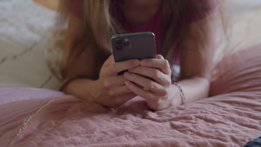 A girl wearing a pink shirt is lying on her stomach on a bed with a pink quilted blanket holding a dark smartphone in both hands