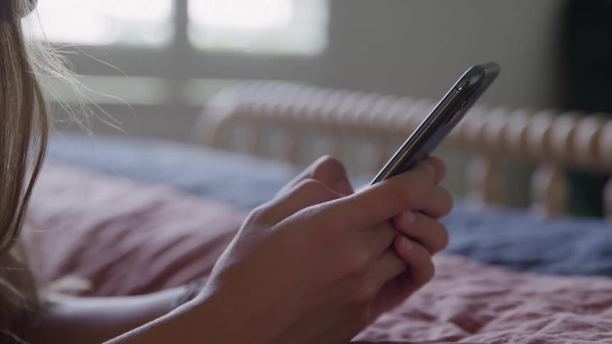 The hands of a girl are seen in a close view holding a black cellphone while lying in bed with light coming from a window in the background