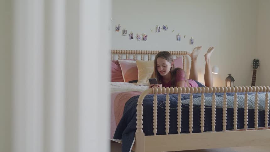 A young girl in a pink shirt is lying on her stomach on a bed with a wooden headboard and colorful covers looking down at her phone with her feet bent up in a bright bedroom