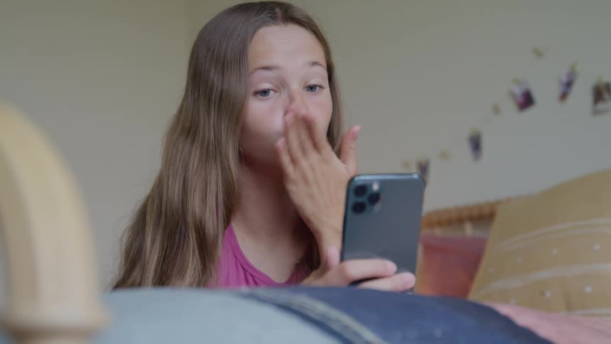 A close view of a young girl with long brown hair wearing a pink shirt as she smiles laughs and waves at a phone during a video call while sitting in a bright bedroom