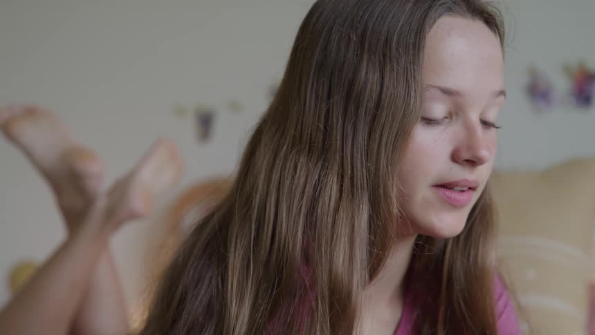 A close view of a young woman with long brown hair wearing a pink shirt as she smiles laughs and waves during a video call while resting in a bright bedroom