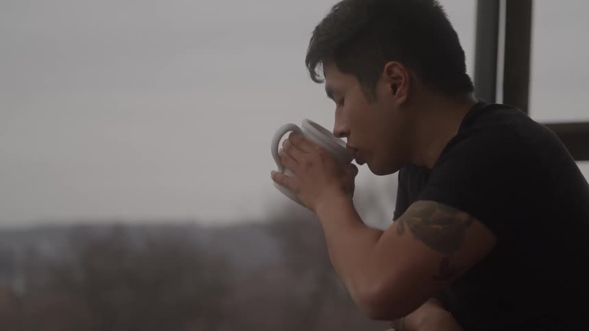 A man with visible tattoos wearing a black shirt stands on a balcony holding a white mug and taking a drink on a foggy cold day with a blurry gray background