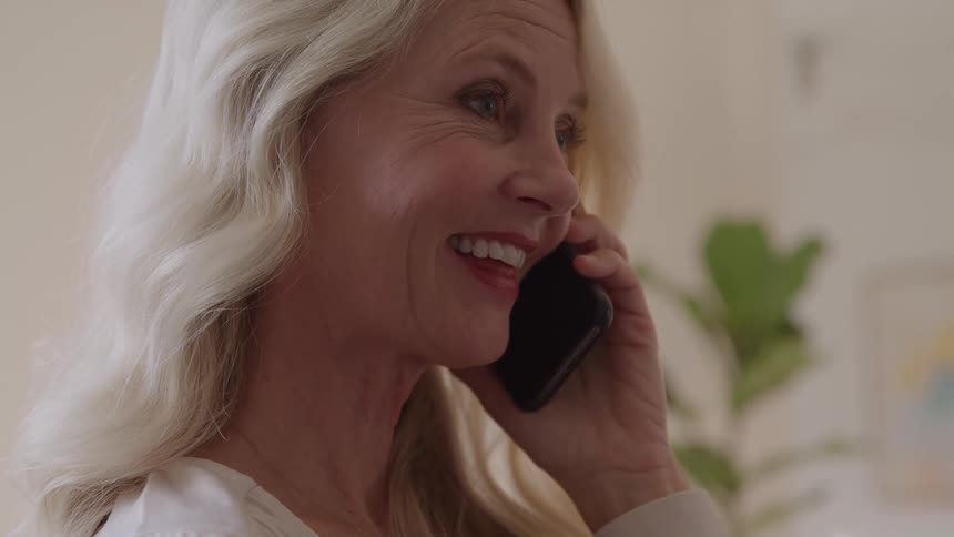 Older woman with light wavy hair smiles while talking on the phone in cozy indoor setting with plant in the background