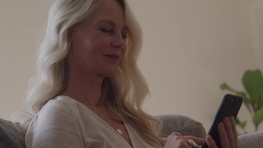 A young woman with blonde hair sits on a couch at midday smiling as she scrolls through her mobile device sunlight filters into the room highlighting a flower plant positioned beside the wall