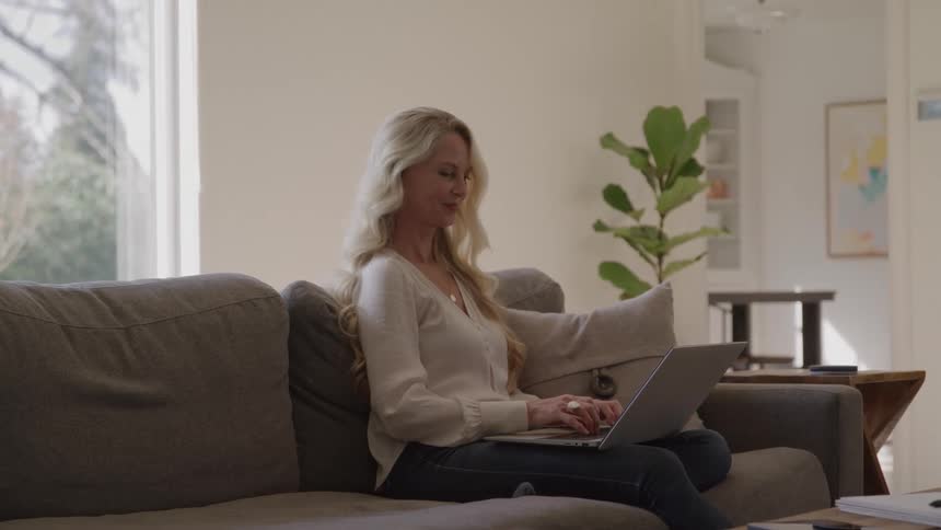 Blondehaired woman uses laptop seated on gray couch in welllit room with plant and shelving in the background