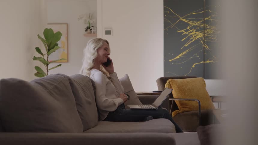 Person sits on gray couch talking on phone while using laptop in modern room with yellow armchair plant and abstract painting