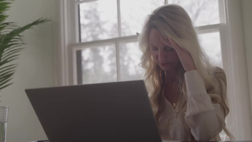 A young woman with blonde hair sits in front of a laptop at midday holding her head in her hands with a sad expression as sunlight filters into the quiet room