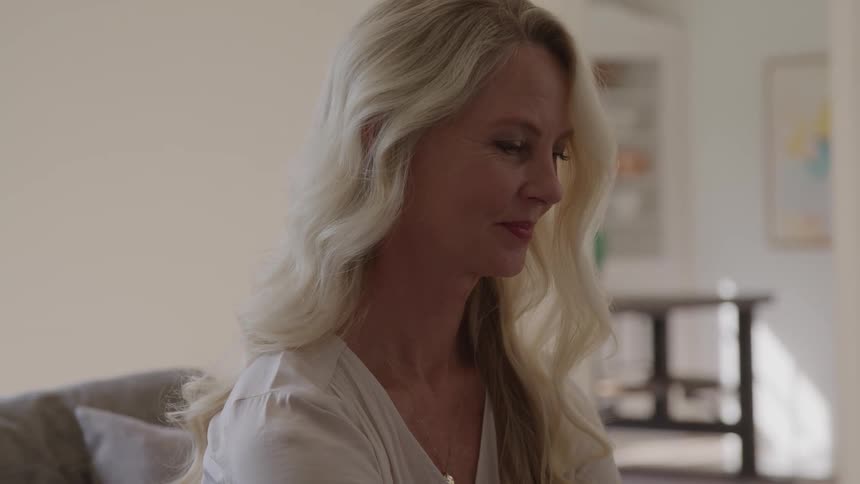A close view of an older woman with long flowing blonde hair wearing a white shirt as she sits with a serious expression in a bright living room