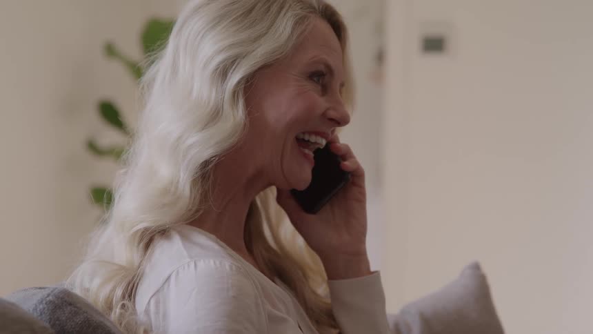 A smiling woman talks on a smartphone in a bright living room