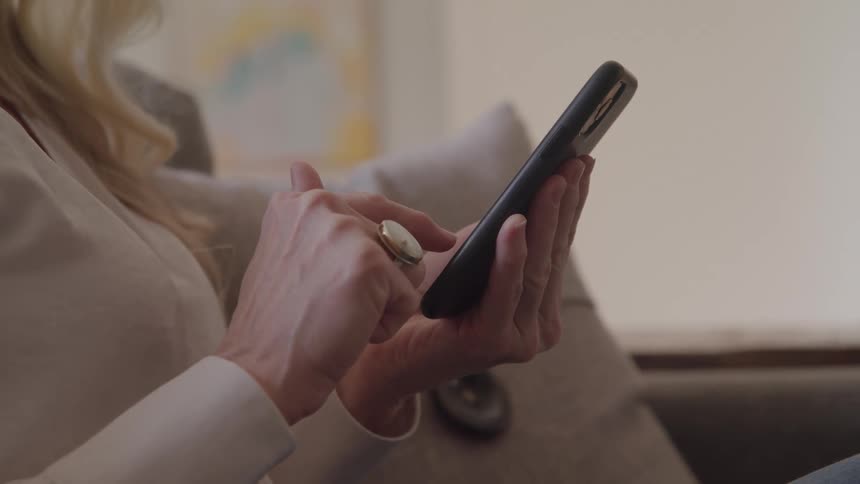 A closeup shot shows a persons hands holding and scrollingtapping on a black smartphone while the person sits on a sofa wearing a lightcolored blazer