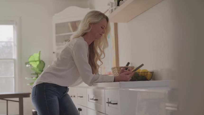 A blonde woman in a white shirt and blue jeans leans on a white kitchen counter while looking down at a black smartphone in a brightly lit kitchen