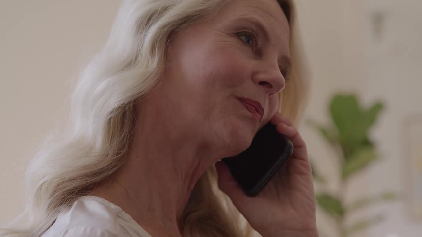 A close view of an older woman with long white hair smiling while she talks on a black smartphone in a bright indoor area with a houseplant nearby