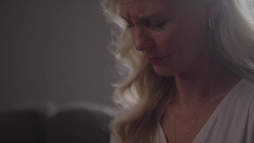 A close side view shows a woman with long wavy blonde hair wearing a white top and a simple gold necklace looking down with a serious and slightly sad expression in a dimly lit room