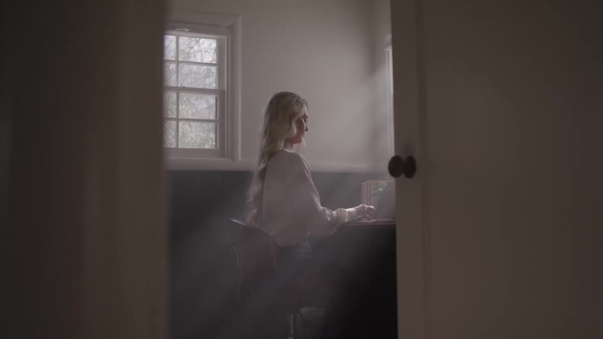 Woman sits in front of a laptop in a room lit by sunlight streaming through the window behind