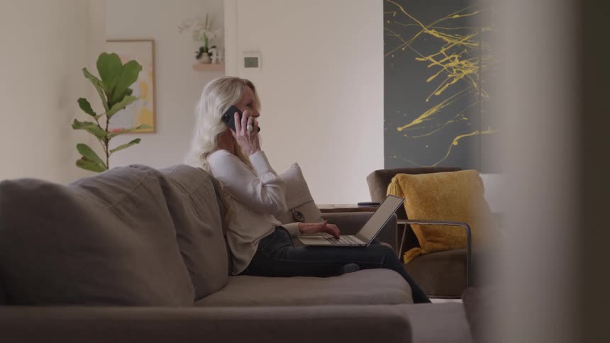 A side medium view of a woman with long blonde hair wearing a white shirt and jeans sitting on a sofa and talking on a cell phone while working on a laptop in a bright modern living room