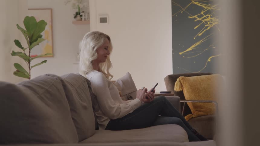 Blonde woman sits on gray couch while interacting with smartphone in room decorated with abstract painting and plant