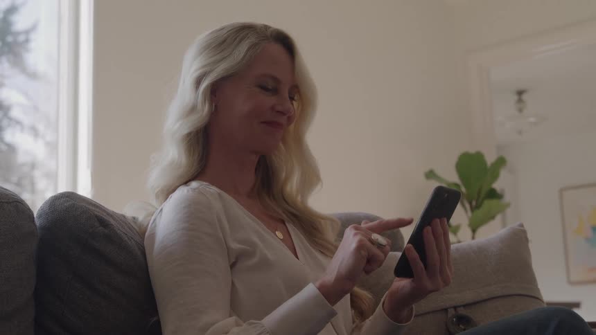 A woman scrolls on her phone while sitting on a sofa in a bright home
