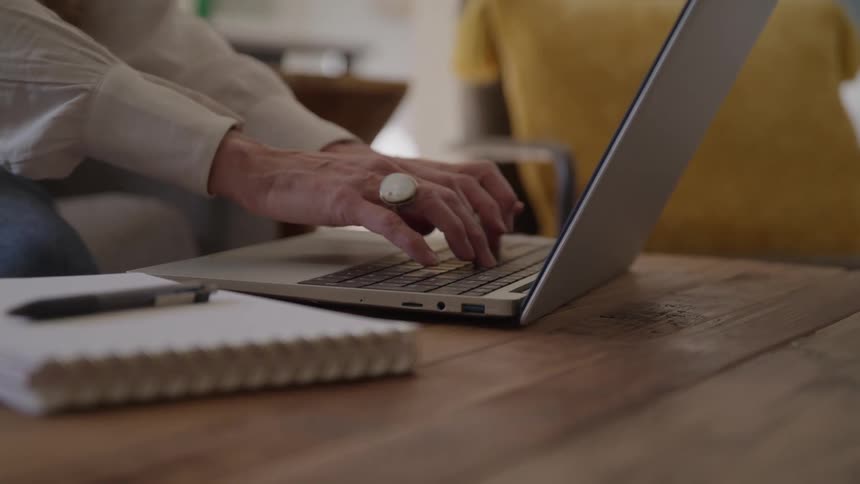 The hands of a woman wearing a white ring are seen typing on a silver laptop next to a notebook and pen on a wooden table in an indoor setting