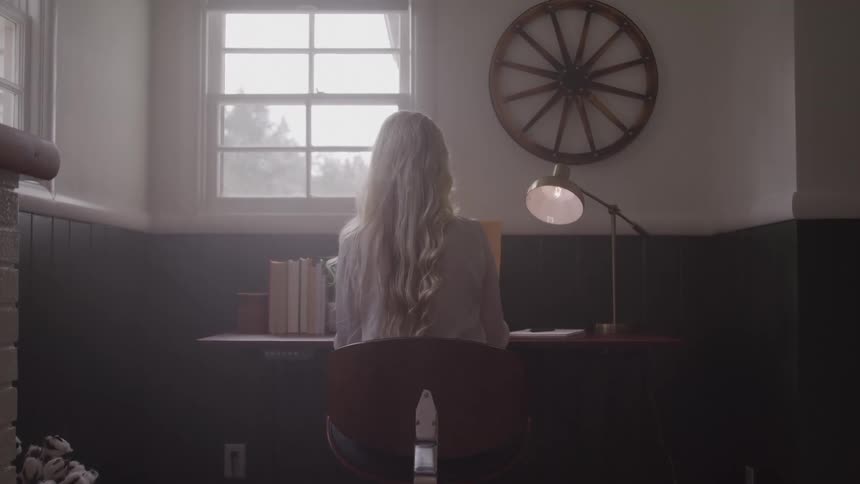 Lighthaired woman sits at desk with books and papers in dim room lit by window