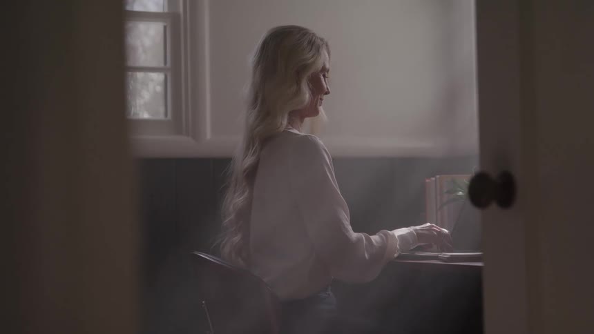 A young blonde woman using a laptop computer while sitting at a desk in a room during daytime