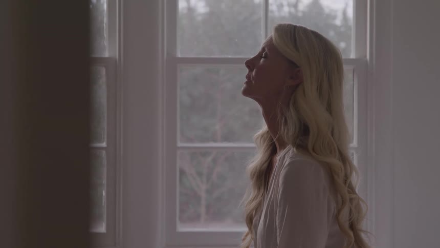 A woman with long blonde hair stands beside a window in her living room at midday she slowly lifts her head from the floor toward the ceiling her expression heavy with sadness as sunlight filters through the glass