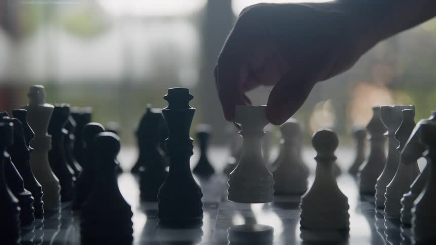 The video is an extreme closeup showing a chessboard set up with black and white pieces a persons hand is visible moving a white piece across the board with a bright hazy background