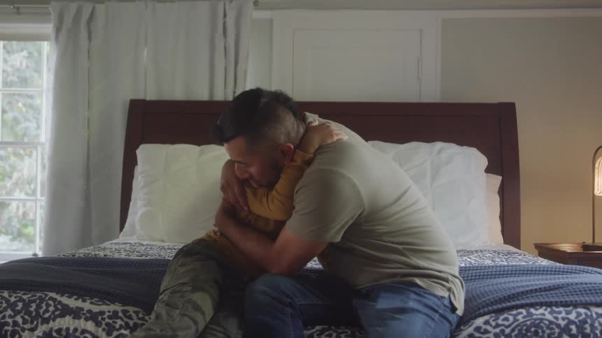 The video shows a man and a young boy sitting together on a bed in a sunlit room smiling and talking the window is visible in the background letting in soft light on the pair as they share an affectionate moment