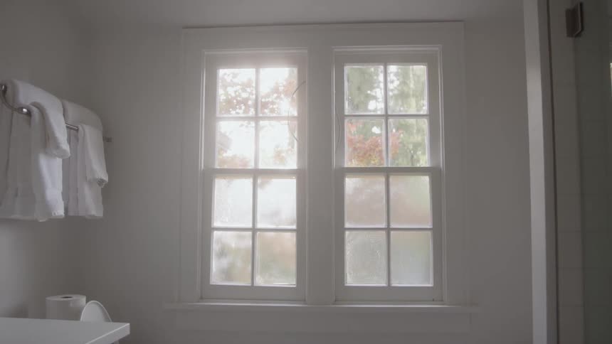 A white bathroom interior focuses on a double window with multiple panes through which bright sunlight shines illuminating the room