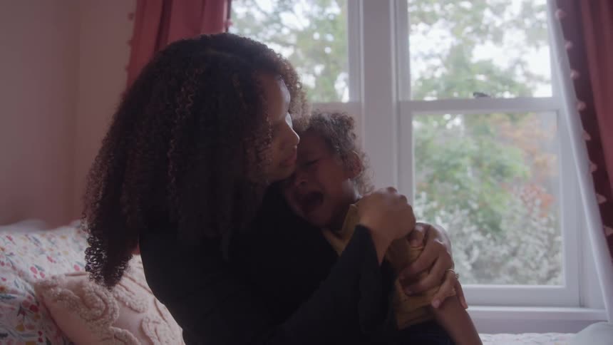A woman with dark curly hair wearing a black shirt holds a small child dressed in a yellow shirt embracing and comforting the crying child near a bright bedroom window