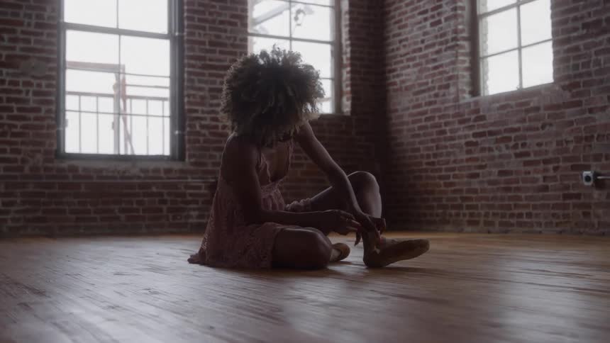 A young woman sits on the floor in an empty room at midday tying her ballet shoe as she prepares to dance
