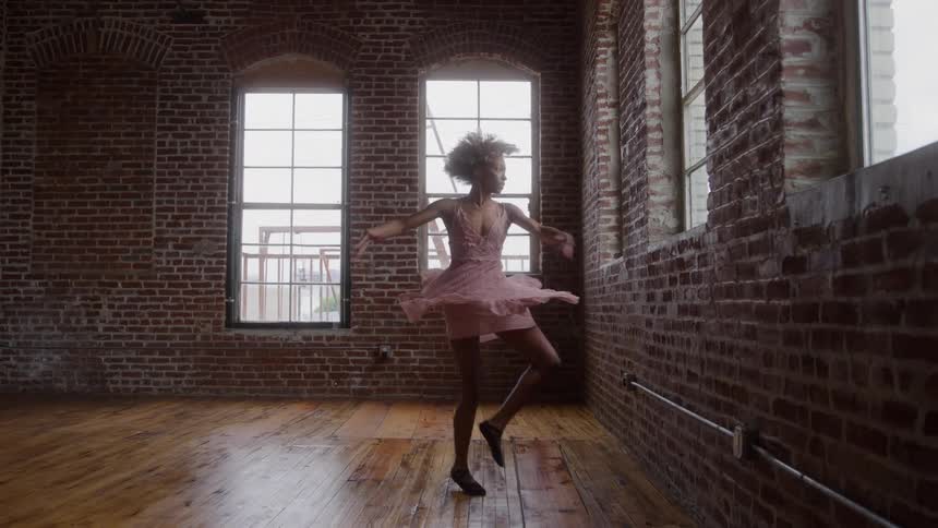A teenage girl dances ballet on a wooden floor at midday wearing a flowing pink dress she moves gracefully inside a spacious room with tall brick walls sunlight streaming through large windows and casting soft patterns across the floor