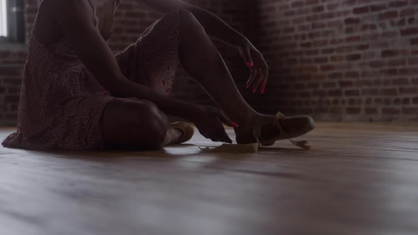 A young female dancer untying and removing her dancing shoes while sitting on the floor in a room during daytime