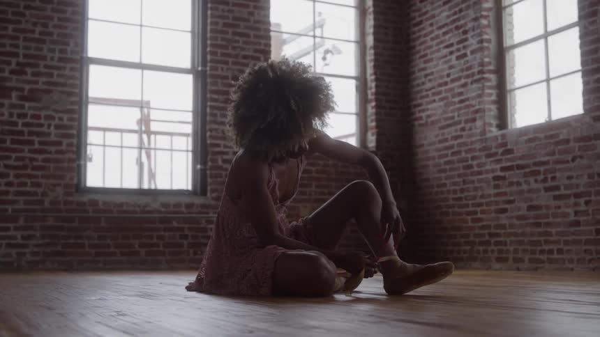 An elegant woman with large natural hair wearing a casual dress sits on a wooden floor in a loft with exposed brick walls adjusting the ankle strap of a ballet slipper against bright light from tall windows