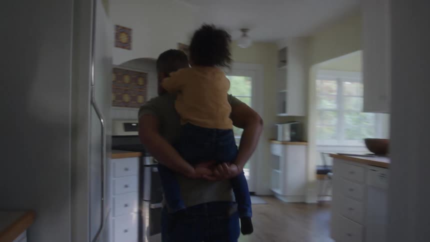 A father playfully carries his daughter on his back while walking around the house at midday both laughing as sunlight filters through the windows filling the space with warmth and joy