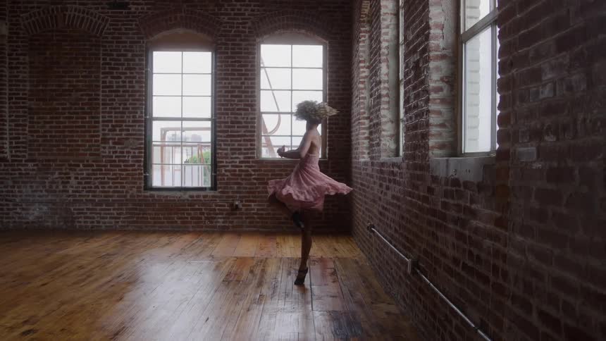 Ballerina in a pink dress spins gracefully in a room lit by tall windows