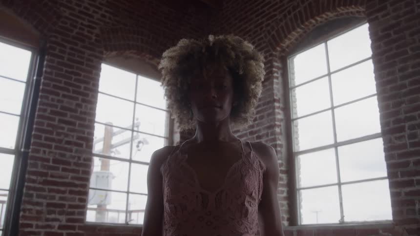Curlyhaired woman in pink lace top stands before arched windows in room with brick wall