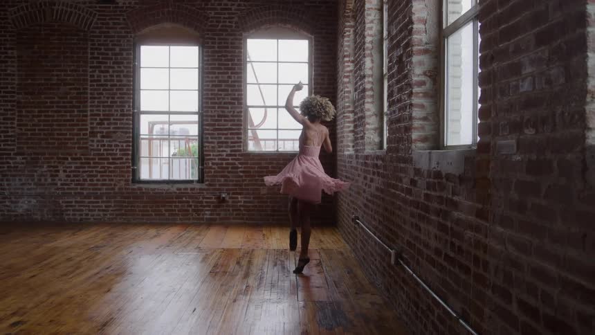A woman in a pink dress gracefully dances ballet in a spacious sunlit industrial loft with exposed brick walls