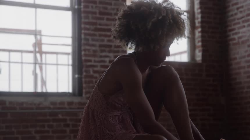 A young woman sits on the floor in an empty room tying her ballet shoe with quiet focus as soft light fills the space