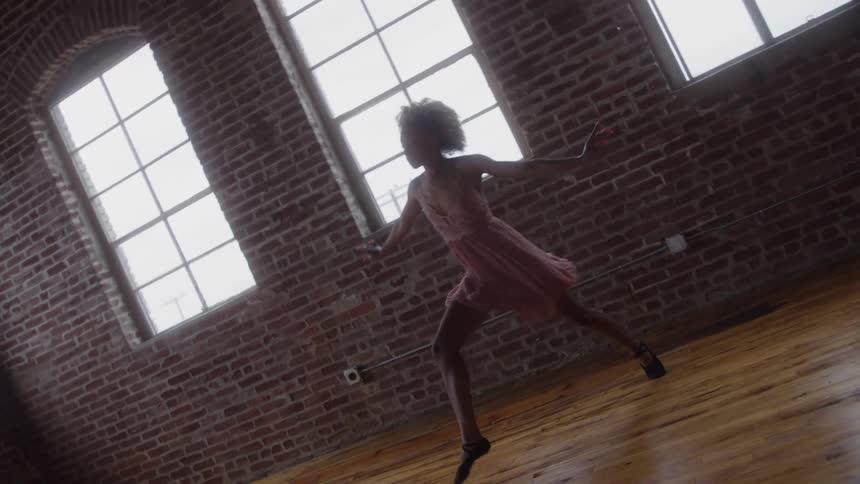 Send a woman with curly hair wearing a pale pink dress and dark shoes dances or leaps across the wooden floor of an empty industrial room with brick walls and large bright windows