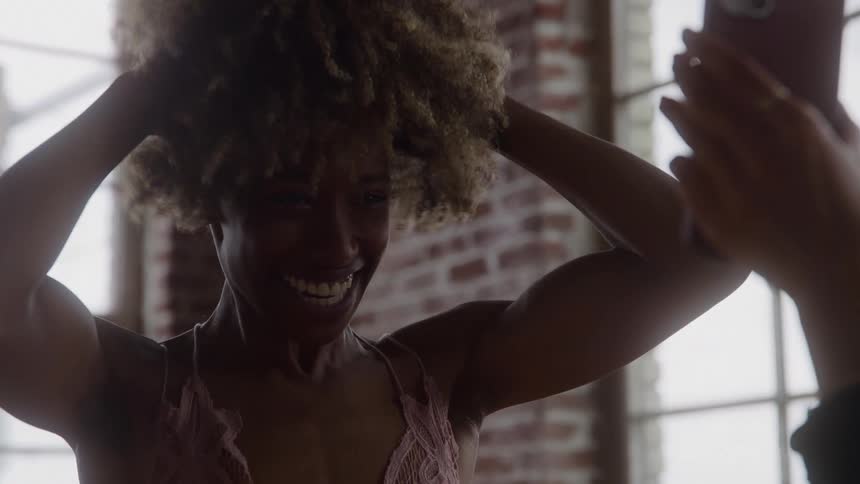 A woman smiling and touching her hair looking at a phone held by another person in front of her