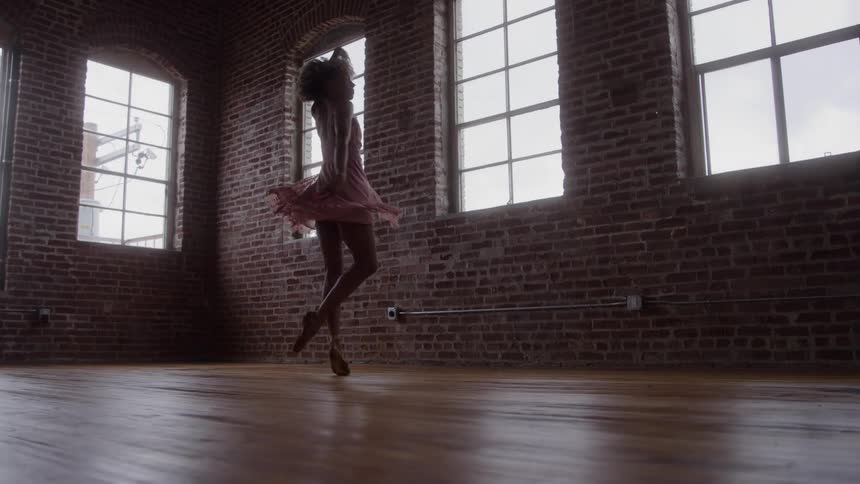 A woman with dark curly hair wearing a pale pink dress is captured dancing with her arm raised while spinning on a shiny wooden floor in a dimly lit room with exposed brick walls and large bright windows