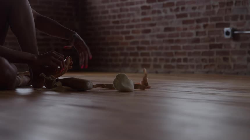 A dancer sits on a polished wood floor near a brick wall preparing for practice by tying the ribbons on a pair of pointe shoes