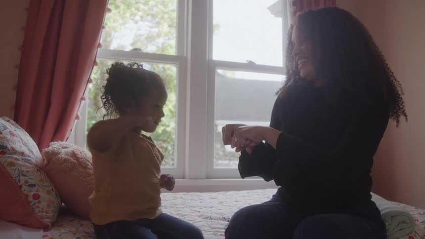 A mother and child sit together on a bed in their sunlit bedroom at midday sharing a joyful playful moment their faces glow with smiles and laughter as they highfive and exchange fist bumps filling the room with warmth and connection