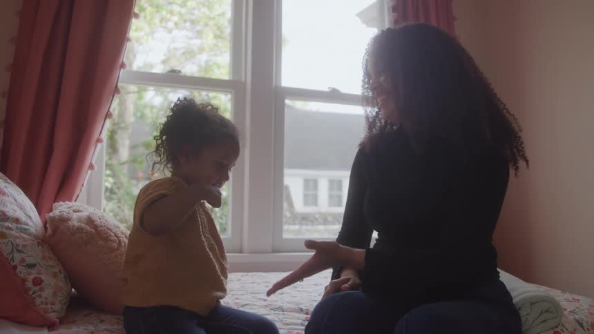 Mother in dark top highfives daughter in yellow shirt sitting on bed with floral blanket in front of large windows