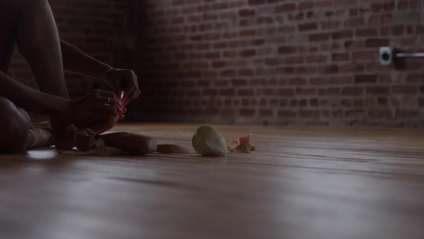 Person ties ballet shoe ribbons seated on wooden floor in room with exposed brick wall